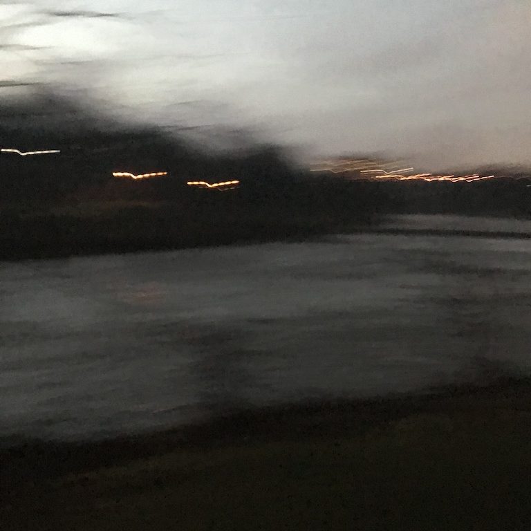 A blurry photo of the river at dusk in dark greys and blues, with lights across the river smeared.